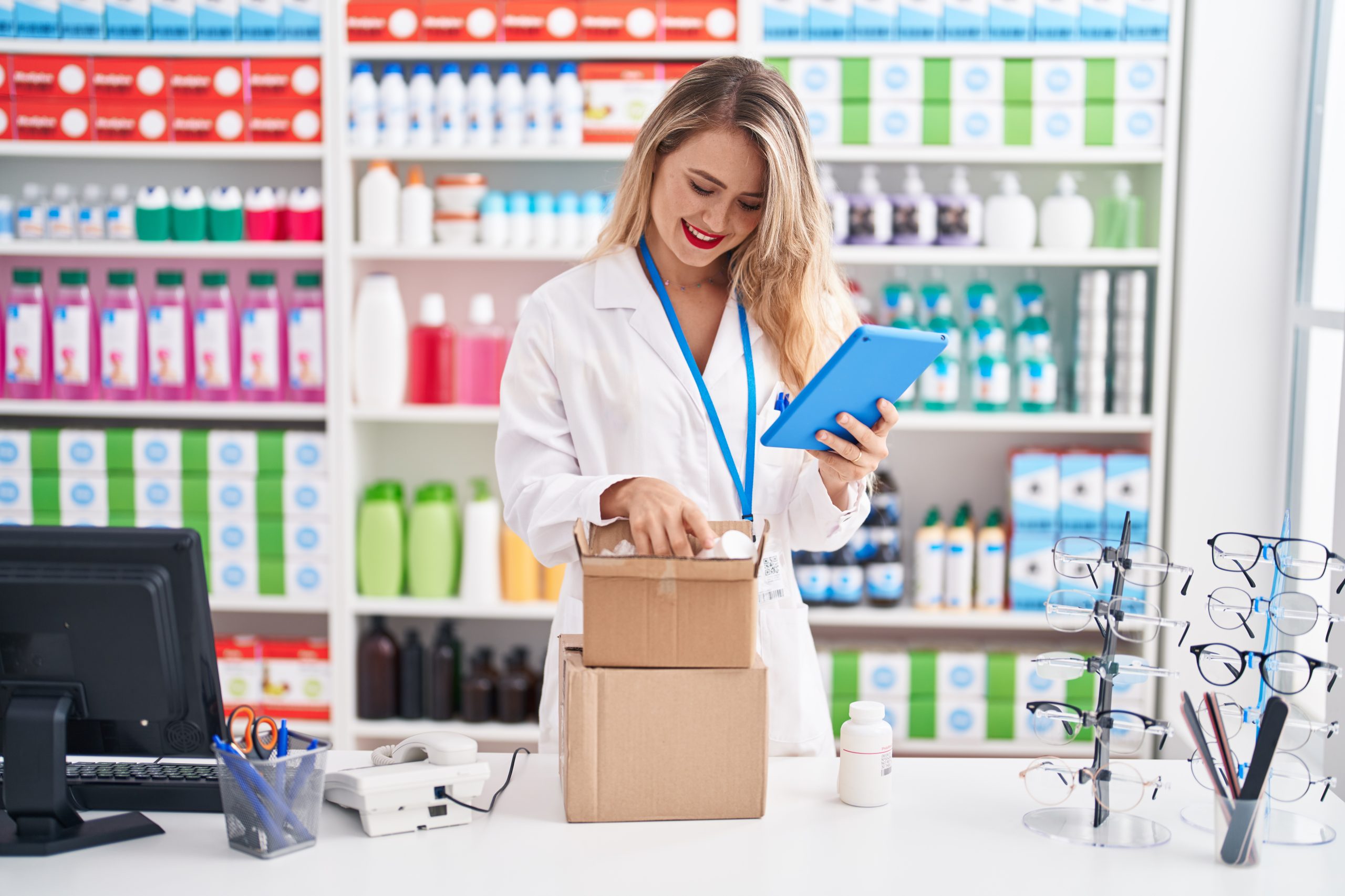 Young beautiful hispanic woman pharmacist using touchpad opening package at pharmacy